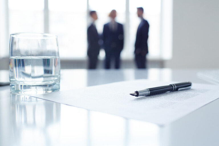 Image of business document, pen and glass of water at workplace with group of colleagues on background